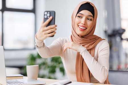 beautiful young muslim woman makes selfie, photographed using smartphone while sitting in office at workplaceの写真素材