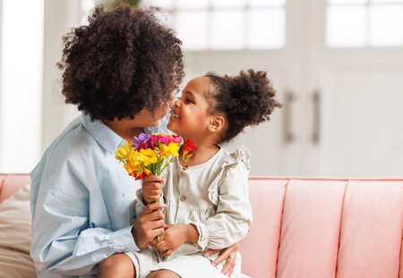 Happy mother's day! Afro american family happy baby daughter congratulations mom on the holiday, hugs her and gives bouquet of flowers at homeの写真素材