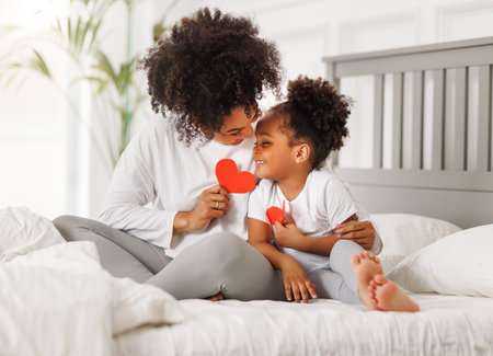 Cute little girl daughter congratulating his mom happy african american woman with Mother's day, giving her handmade greeting postcard with red heart at home. family holidays conceptの写真素材
