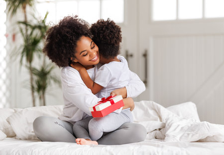 happy mother's day. african american baby daughter giving gift box to her mom for holiday in bed at homeの写真素材