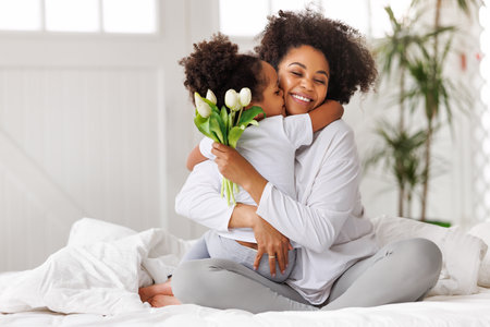 Happy mother's day! Afro american family happy baby daughter congratulations mom on the holiday, hugs her and gives bouquet of flowers at homeの写真素材