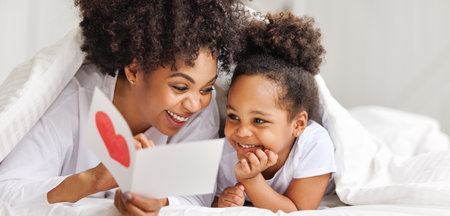 Cute little girl daughter congratulating his mom happy african american woman with Mother's day, giving her handmade greeting postcard with red heart at home. family holidays conceptの写真素材