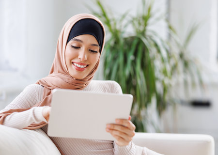 beautiful young smiling muslim woman in traditional religious hijab uses tablet while sitting on sofa at homeの写真素材