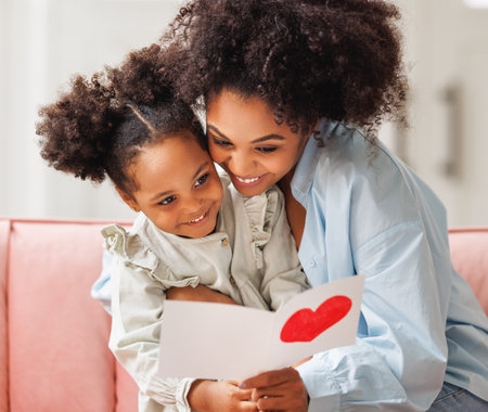 Cute little girl daughter congratulating his mom happy african american woman with Mother's day, giving her handmade greeting postcard with red heart at home. family holidays conceptの写真素材