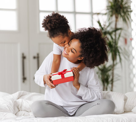 happy mother's day. african american baby daughter giving gift box to her mom for holiday in bed at homeの写真素材