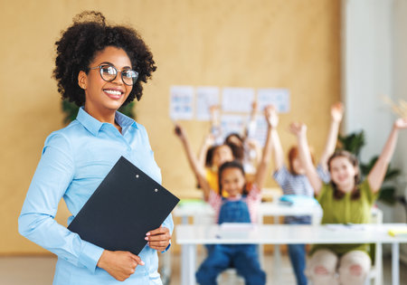 Portrait of friendly african american woman teacher smiling and standing in bright classroom during lesson with kids at schoolの写真素材