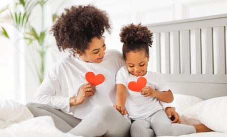 Cute little girl daughter congratulating his mom happy african american woman with Mother's day, giving her handmade greeting postcard with red heart at home. family holidays conceptの写真素材