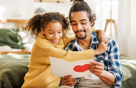 Happy african american father receiving gift for fathers day from kid daughter, cute curly girl giving handmade greeting card with hand drawn heart to dad for birthday while sitting on bed at homeの写真素材