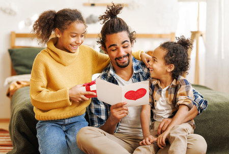 Happy african american father receiving gift for fathers day from kids daughter and son, children giving handmade greeting card with hand drawn heart to dad for birthday while sitting on bed at homeの写真素材