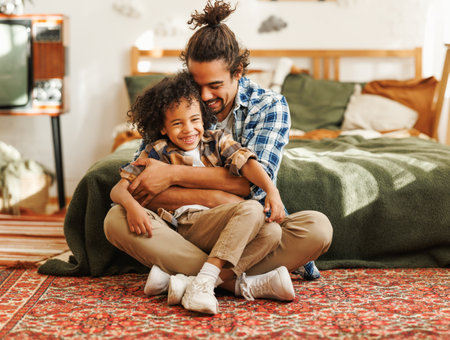 Happy afro american family father and son laughing and playing while sitting on bed in cozy bedroom at home and enjoying fathers day together, young loving dad tickling embracing child little boyの写真素材