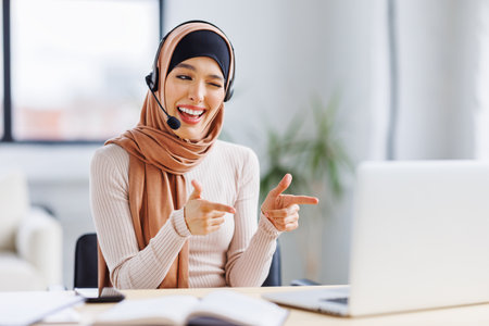 friendly smiling muslim woman in traditional dress and with headset smiling and gesticulating while sitting at desk in home office and talking to online coworkers during online meetingの写真素材