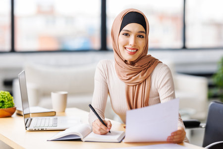 beautiful young smiling muslim woman in traditional religious hijab works remotely on laptop from homeの写真素材