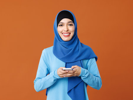 Beautiful overjoyed young smiling muslim woman in traditional religious hijab reading great news on smartphone, amazed young muslim lady receiving unexpected message on mobile phone, isolated on orange studio backgroundの写真素材