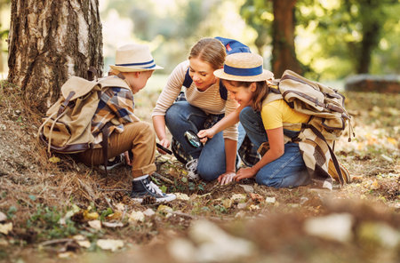 Happy family: two kids boy and girl with backpacks looking examining environment through magnifying glass while exploring forest nature on sunny day during outdoor ecology school lessonの写真素材