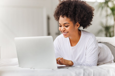 happy young african american woman relaxing with laptop at home lying in bed in bright bedroomの写真素材