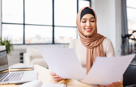 beautiful young smiling muslim woman in traditional religious hijab works remotely on laptop from homeの写真素材