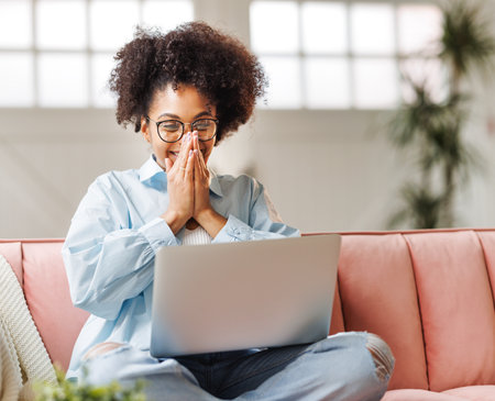 beautiful young smiling african american woman smiling and rejoices in victory sitting on sofa and working at laptop screen after finishing project in home officeの写真素材