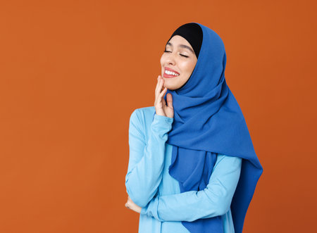 portrait of beautiful young smiling muslim woman in traditional religious hijab on orange backgroundの写真素材