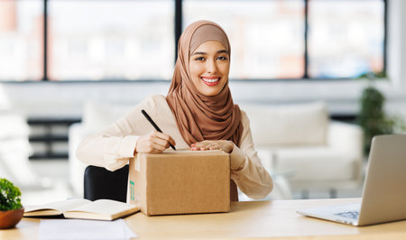 Beautiful young smiling muslim woman in traditional religious hijab signs carton box to send parcel while at table in home officeの写真素材
