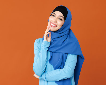 portrait of beautiful young smiling muslim woman in traditional religious hijab on orange backgroundの写真素材