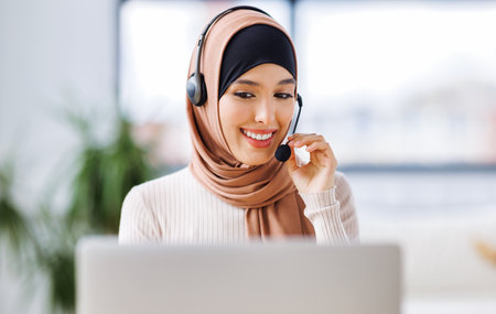 friendly smiling muslim woman in traditional dress and with headset working in front of laptop talking while calling clientの写真素材