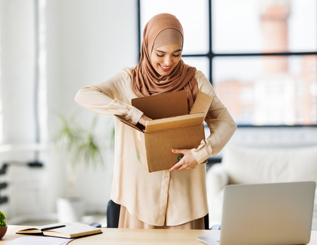 Beautiful young smiling muslim woman in traditional religious hijab unpacking delivered order from cardboard box while at table in home officeの写真素材