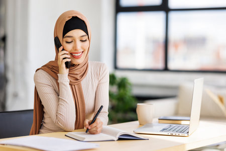 Friendly smiling Muslim woman smiling and making call via smartphone while working remotely from home while sitting at desk and talking to coworkers. Aerobic woman freelancer talking on mobile phone during remote workの写真素材