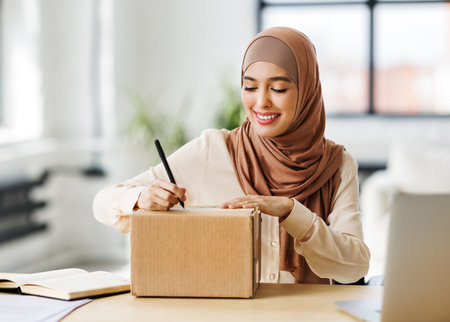 Beautiful young smiling muslim woman in traditional religious hijab signs carton box to send parcel while at table in home officeの写真素材