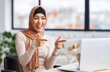 beautiful young smiling muslim woman in traditional religious hijab working remotely behind a laptop, communicates by video call at homeの写真素材