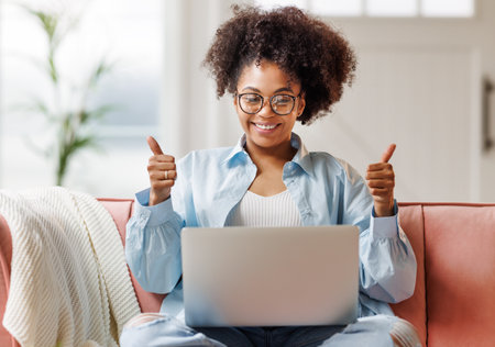 beautiful young smiling african american woman with curly hair uses laptop smiling, gesticulating and talking to online while sitting on sofa at homeの写真素材