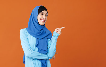 portrait of beautiful young smiling muslim woman in traditional religious hijab pointing to side on orange backgroundの写真素材