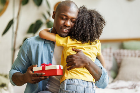 Happy african american family father with excited child son hugging smiling while celebrating Fathers day together at home. Boy congratulating giving dad gift boxの写真素材