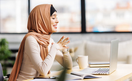 beautiful young smiling muslim woman in traditional religious hijab working remotely behind a laptop, communicates by video call at homeの写真素材