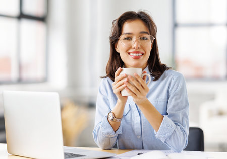 beautiful young smiling woman relaxing with cup of hot drink during break from work working remotely behind a laptop at homeの写真素材
