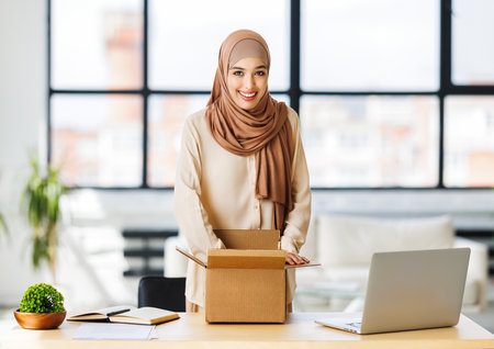 Beautiful young smiling muslim woman in traditional religious hijab unpacking delivered order from cardboard box while at table in home officeの写真素材