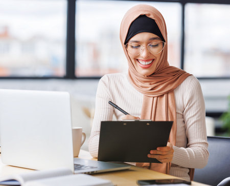 beautiful young smiling muslim woman in traditional religious hijab works remotely on laptop from homeの写真素材