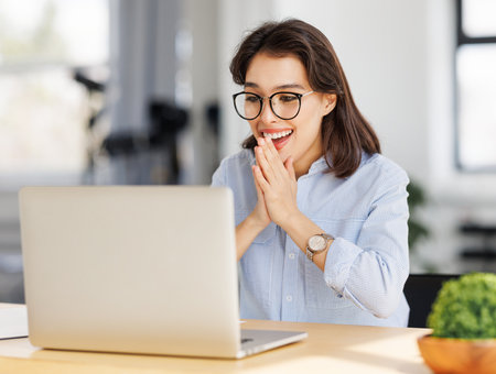 Joyful young business woman freelancer entrepreneur in glasses smiling and rejoices in victory while sitting at desk and working at laptop screen after finishing project in home officeの写真素材