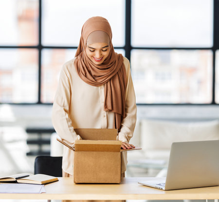 Beautiful young smiling muslim woman in traditional religious hijab unpacking delivered order from cardboard box while at table in home officeの写真素材