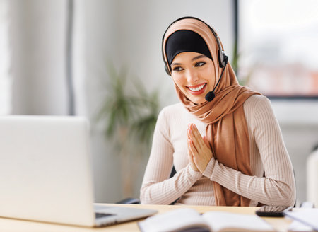 friendly smiling muslim woman in traditional dress and with headset smiling and gesticulating while sitting at desk in home office and talking to online coworkers during online meetingの写真素材