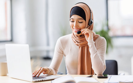 friendly smiling muslim woman in traditional dress and with headset working in front of laptop talking while calling clientの写真素材
