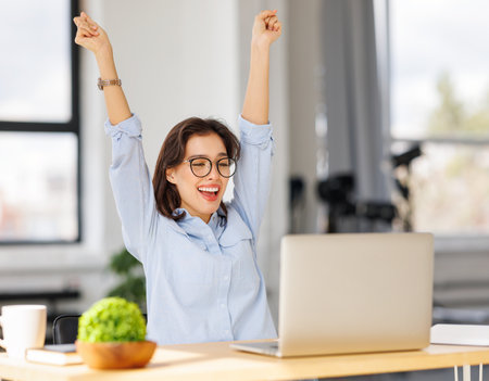 Joyful young business woman freelancer entrepreneur in glasses smiling and rejoices in victory while sitting at desk and working at laptop screen after finishing project in home officeの写真素材