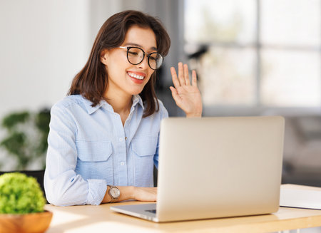 Joyful happy young female entrepreneur in glasses smiling and gesticulating while sitting at desk in home office and talking to online coworkers during online meetingの写真素材