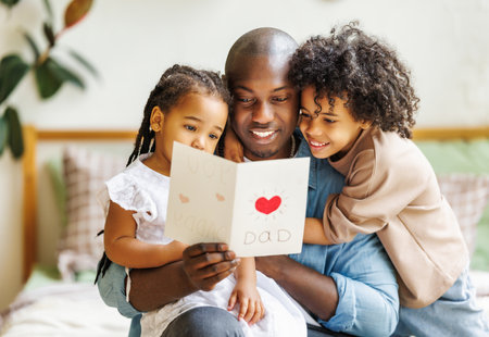 We love you dad. African american happy father getting congratulations on Fathers Day from two excited kids at home, son and daughter, smiling children giving daddy handmade postcardの写真素材