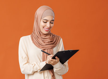 Beautiful woman in traditional muslim hijab writing notes on orange backgroundの写真素材