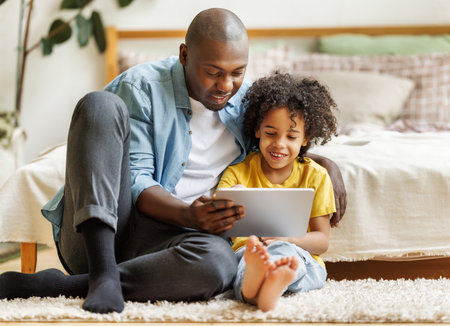 Happy african american family little boy son with young dad watching funny videos on tablet together at home, father with child using computer in bedroom while enjoying weekendの写真素材