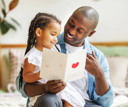 African american happy family father with baby daughter hugging smiling while celebrating Fathers day together at home. Little girl congratulating giving dad homemade greeting postcardの写真素材