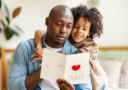 African american happy family father with excited child son hugging smiling while celebrating Fathers day together at home. Little boy congratulating giving dad homemade greeting postcardの写真素材