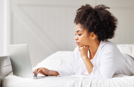happy young african american woman relaxing with laptop at home lying in bed in bright bedroomの写真素材