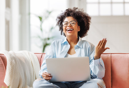 beautiful young smiling african american woman with curly hair uses laptop smiling, gesticulating and talking to online while sitting on sofa at homeの写真素材