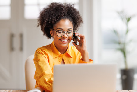 Beautiful smiling african american woman making call via smartphone while working remotely from home while sitting at desk and talking to coworkers. Ethnic female freelancer talking on mobile phone during remote workの写真素材